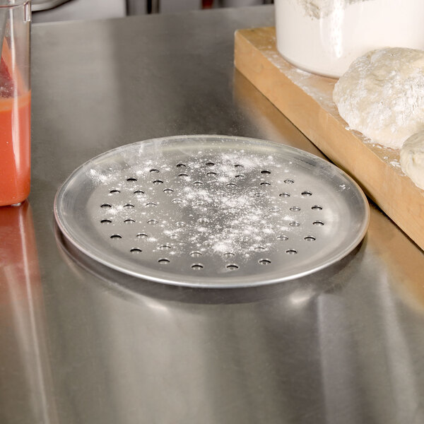 An American Metalcraft perforated aluminum pizza pan with dough on it next to a cutting board.