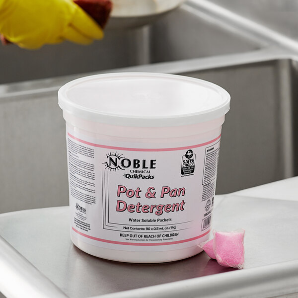 A white container with a pink label of Noble Chemical QuikPacks Concentrated Pot & Pan Detergent on a metal surface.