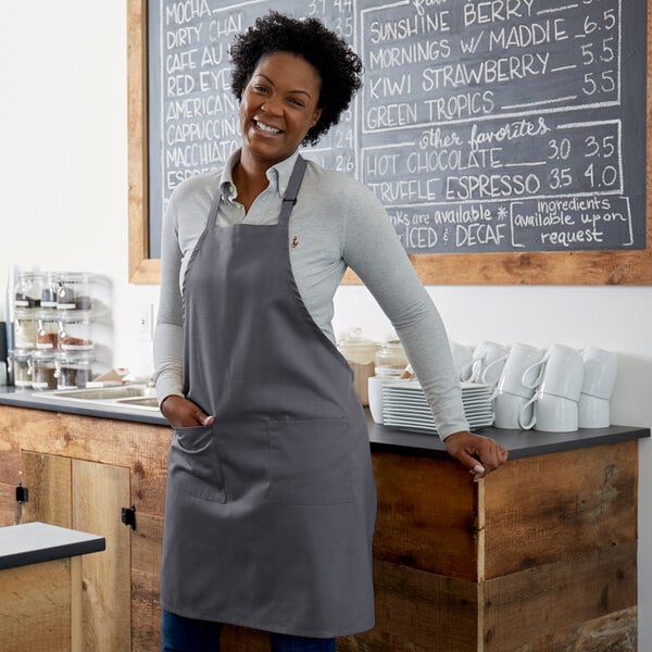 A woman wearing a Choice gray poly-cotton bib apron with 2 pockets standing in a cafe.