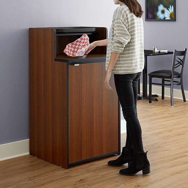 A woman standing in front of a Lancaster Table & Seating walnut trash receptacle enclosure.