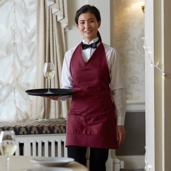 A woman wearing a burgundy tuxedo apron holding a tray with a glass of wine.