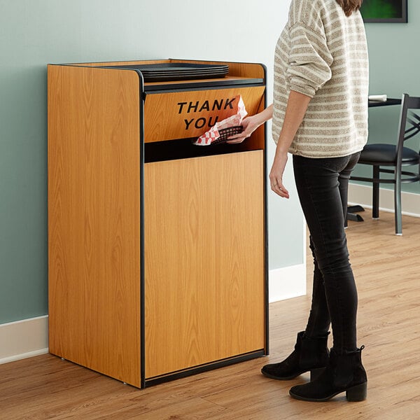 A woman standing next to a Lancaster Table & Seating natural wooden trash can enclosure with a "THANK YOU" swing door.