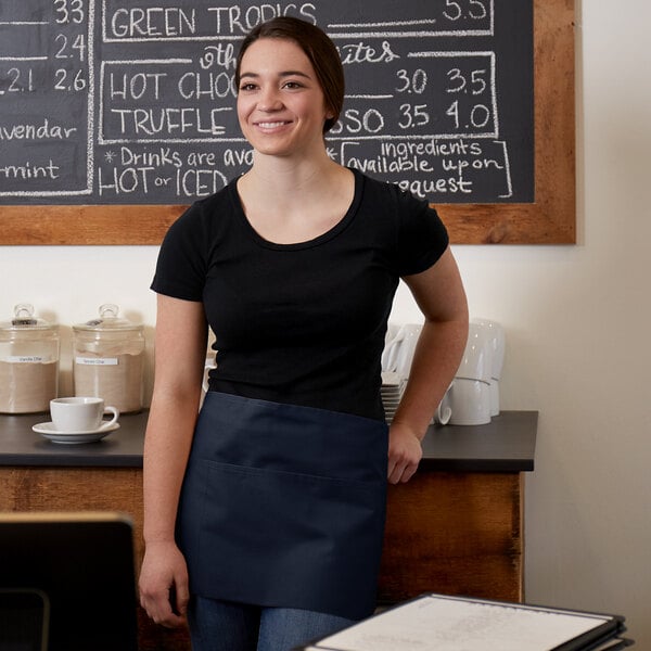 A woman wearing a navy blue standard waist apron with three pockets standing in front of a chalkboard menu.