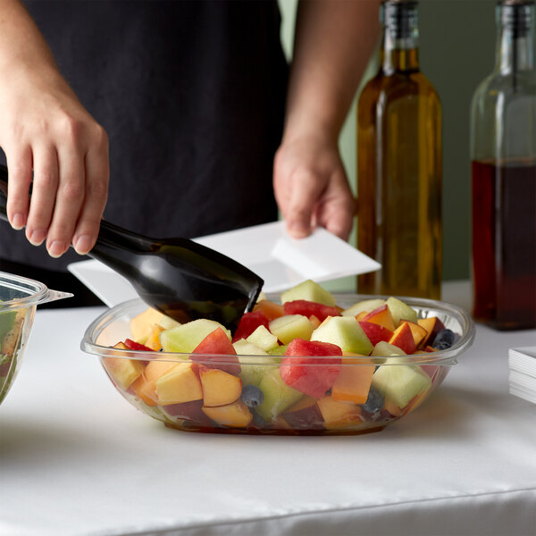 A person pouring fruit into a Fineline clear plastic bowl on a table.