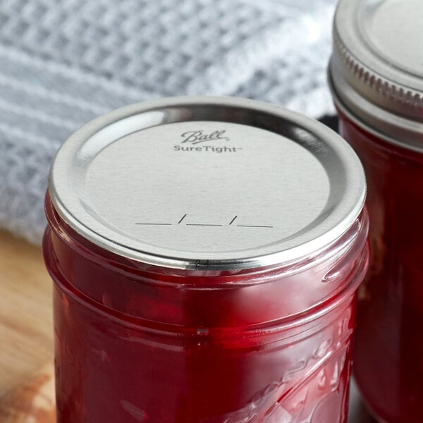 A close-up of two Ball canning jars with red jam inside, each sealed with a Ball Regular Mouth lid.
