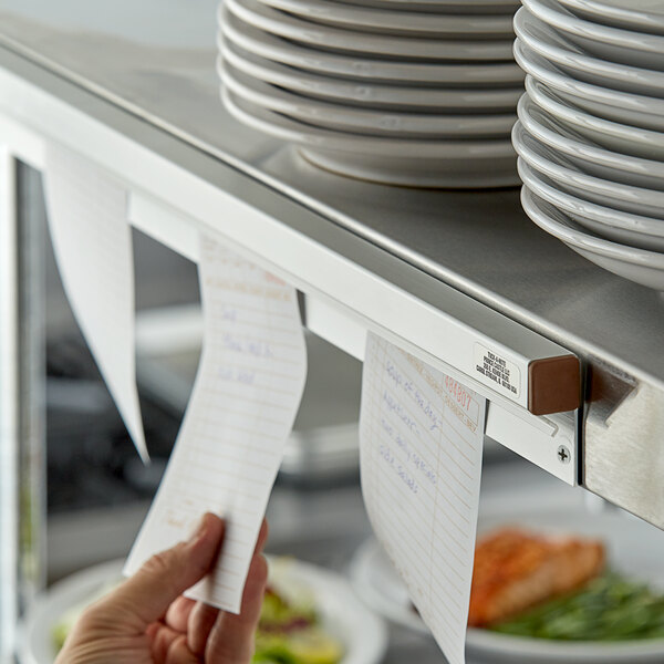 A person using a Prince Castle wall-mounted ticket holder to display a paper with writing on it over a shelf with stacks of plates.