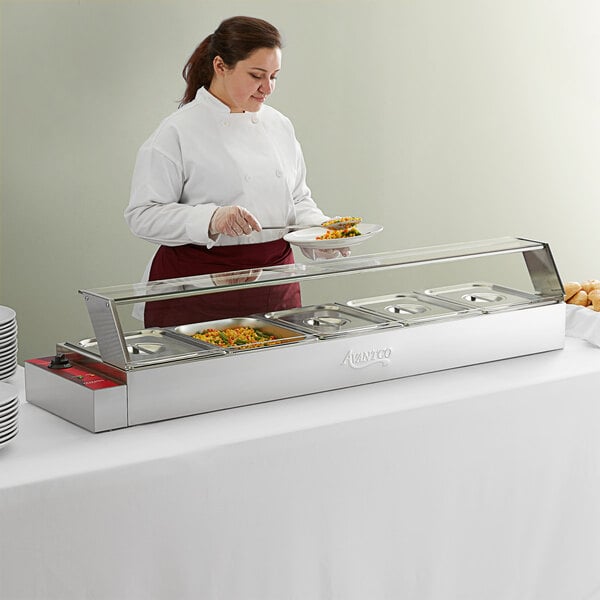 A woman in a white apron serving food from an Avantco countertop food warmer at a buffet.