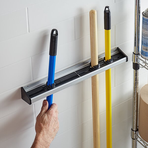 A person using a Prince Castle 918-B Mop and Broom Rack to hold mops and brooms on a wall.