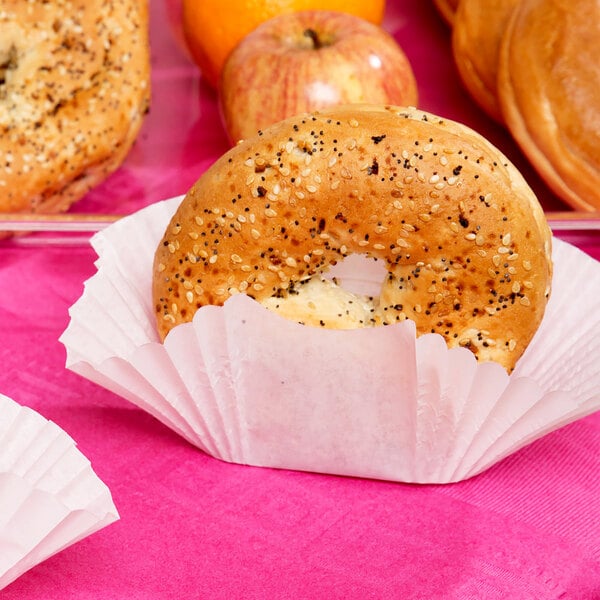 A white paper Hoffmaster fluted bagel cup holding a bagel on a table with apples.