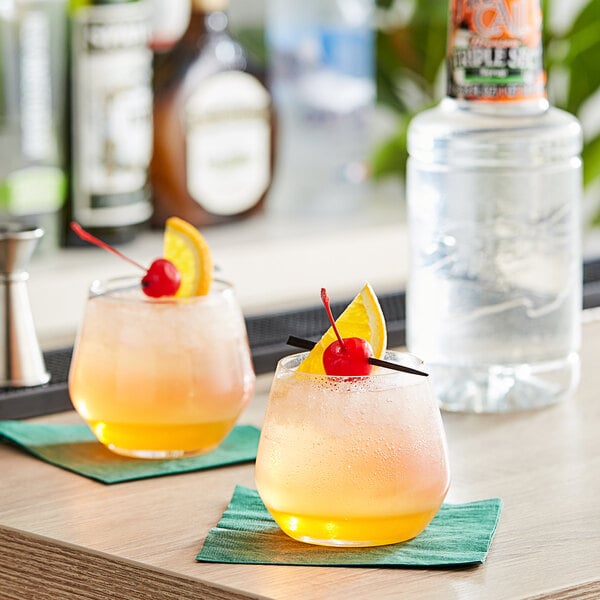 Two glasses of orange drinks with a cherry and a straw sit on a bar counter with a bottle of Finest Call Premium Triple Sec syrup.