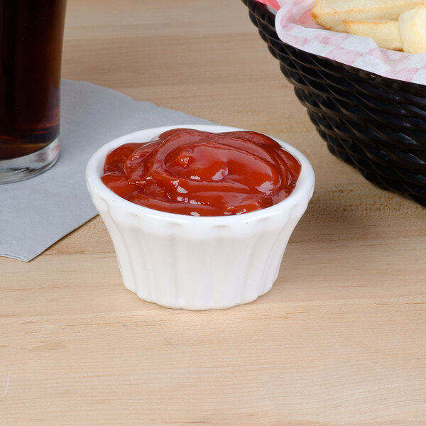 A flared white Hall China ramekin filled with ketchup on a table with a basket of french fries.
