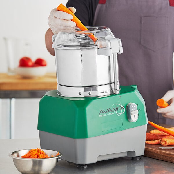 A person using an AvaMix stainless steel food processor to chop a carrot.