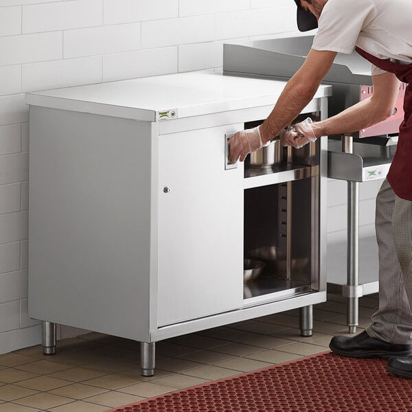 A man in a white shirt and apron opening a stainless steel cabinet.