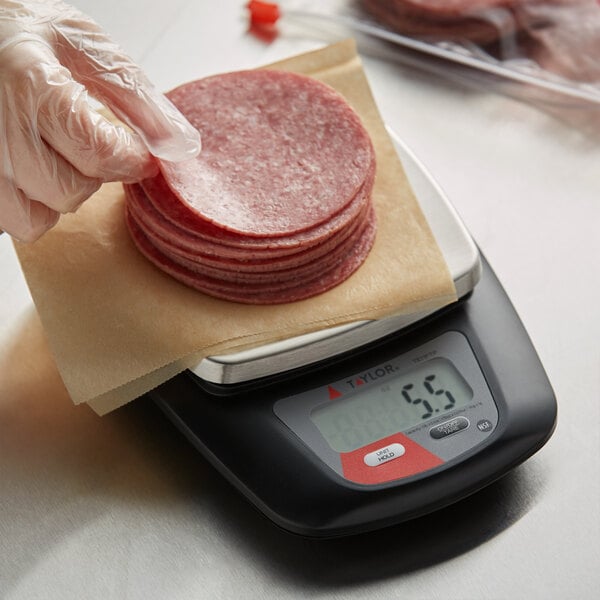 A person wearing a plastic glove weighing meat on a Taylor stainless steel digital portion scale.