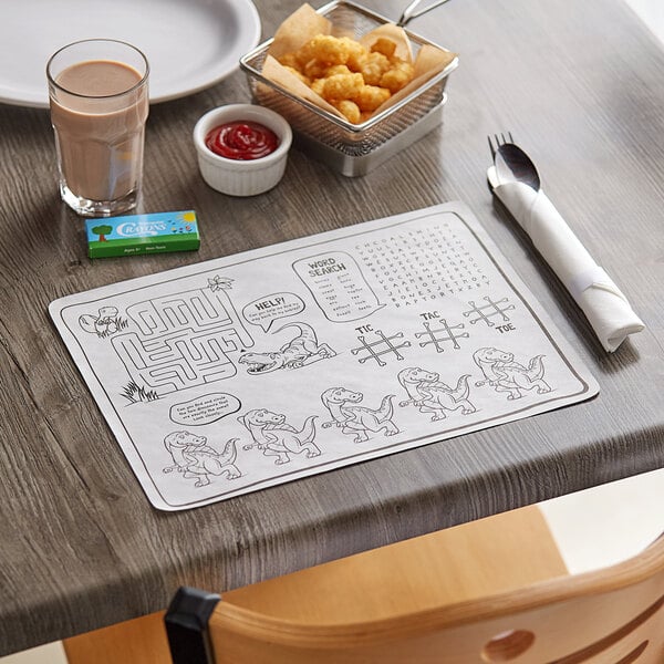 A wood table with a Choice Kids Dinosaur Interactive Place mat, a glass of chocolate milk, and a spoon wrapped in a napkin.