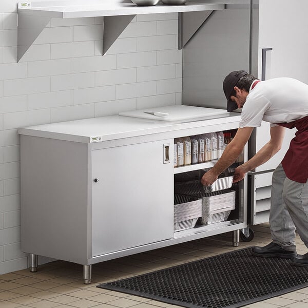 A man in a white shirt and black pants opens the sliding doors on a Regency stainless steel enclosed base table in a professional kitchen.