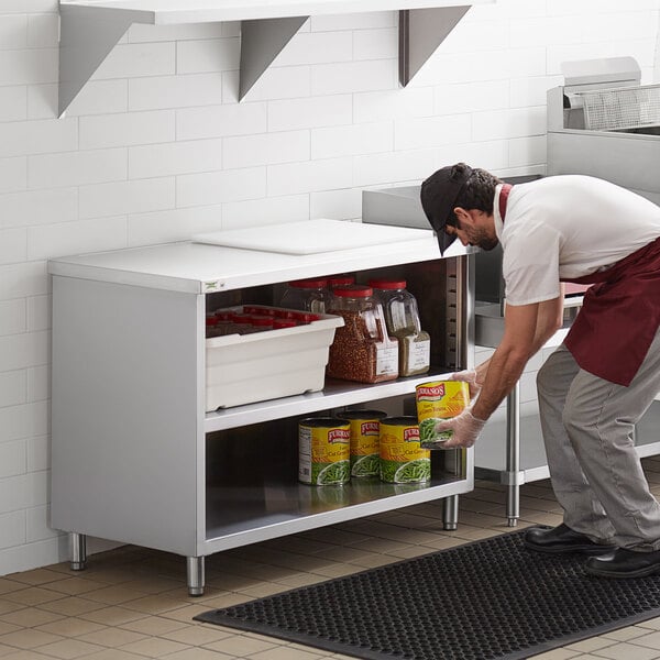 A man in a white shirt and black pants using a Regency stainless steel enclosed base table with a midshelf to store food.