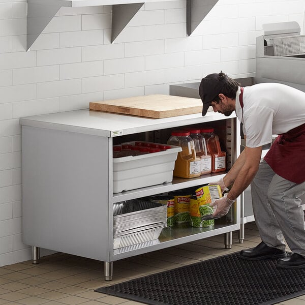 A man in a white shirt and black pants reaching for a shelf on a Regency stainless steel table.