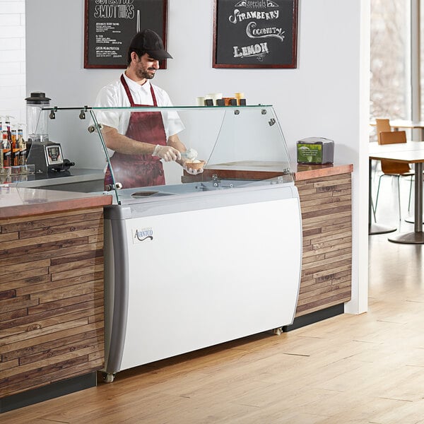 An Avantco ice cream dipping cabinet with a clear top on a counter with a man in a white apron.