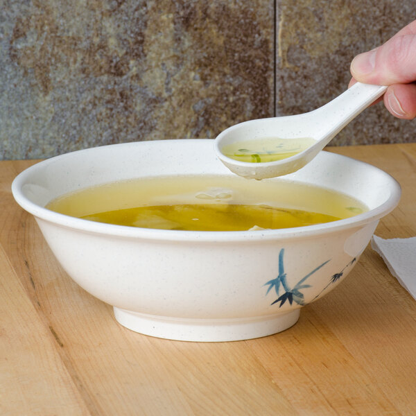 A hand using a Thunder Group Blue Bamboo Chinese soup spoon to pour broth into a bowl.