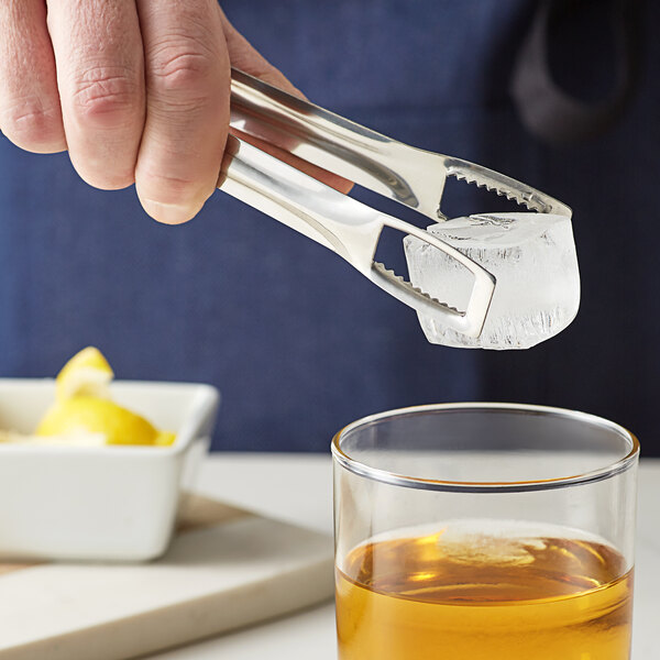 A person using Arcoroc serrated ice tongs to add a lemon peel to a glass of liquid.