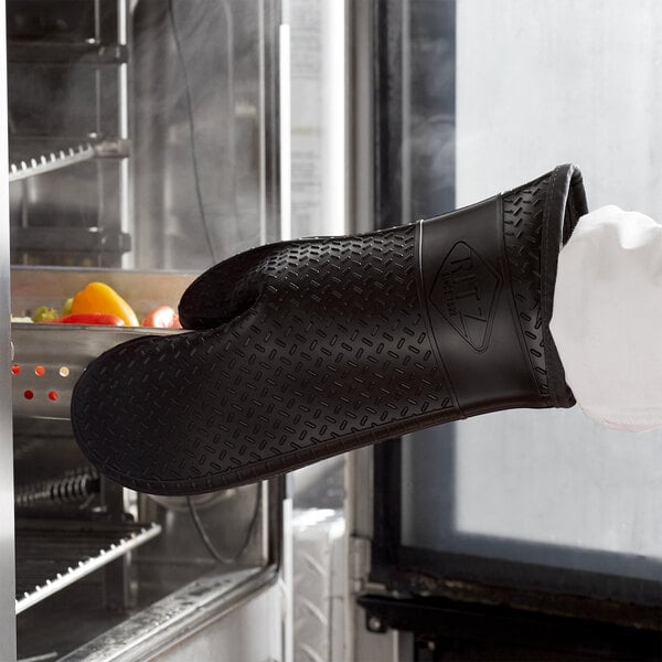 A gloved hand using a black RITZ silicone oven mitt to hold a tray of food.