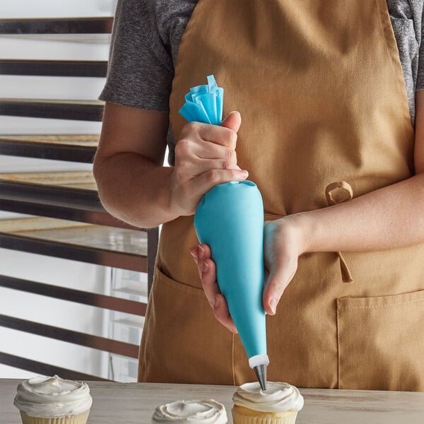 A person in an apron holding a blue Ateco pastry bag over a cupcake.