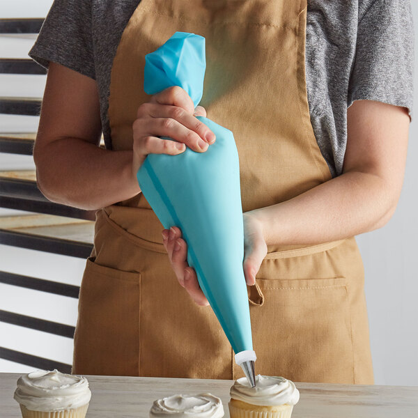 A person in an apron using a blue Ateco pastry bag to frost a cupcake.