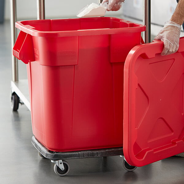 A person wearing plastic gloves to push a Rubbermaid red trash can on a dolly.