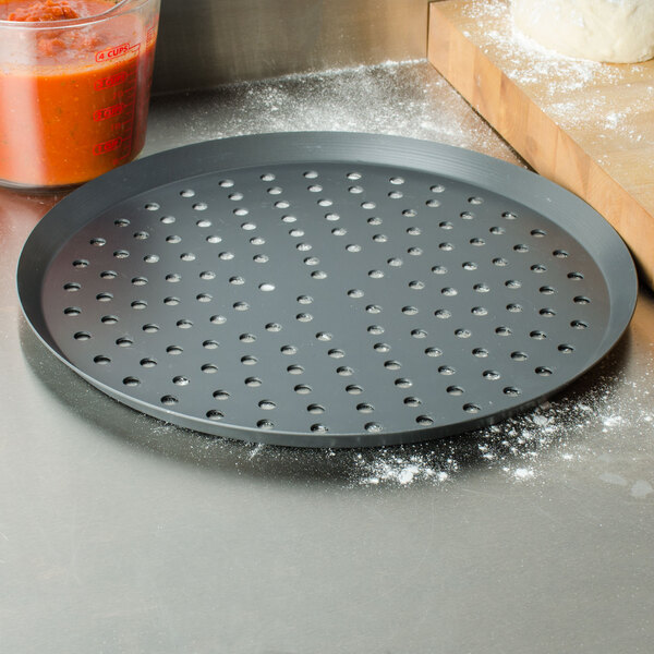 An American Metalcraft black hard coat anodized aluminum pizza pan with perforations on a counter with a pizza.