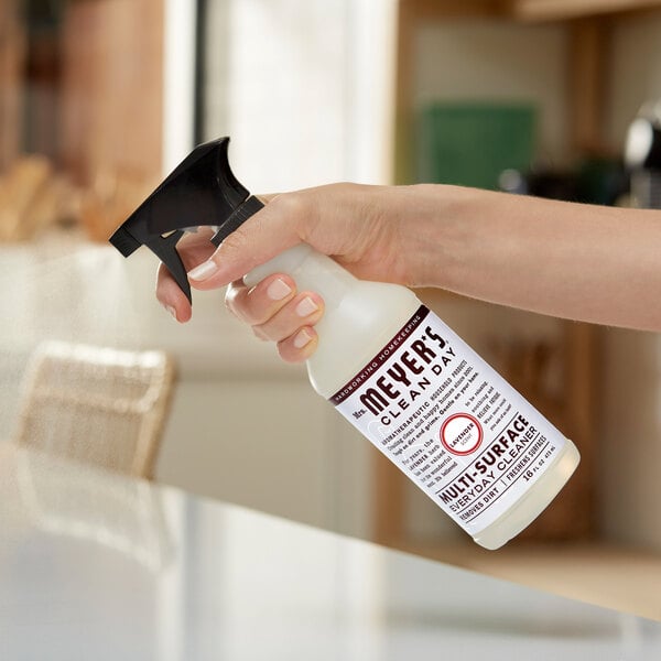 A person holding a white Mrs. Meyer's Clean Day spray bottle on a counter.