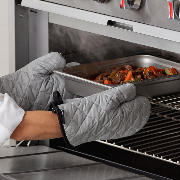 A person wearing San Jamar silicone-coated oven mitts removes a tray of meat and vegetables from an oven.