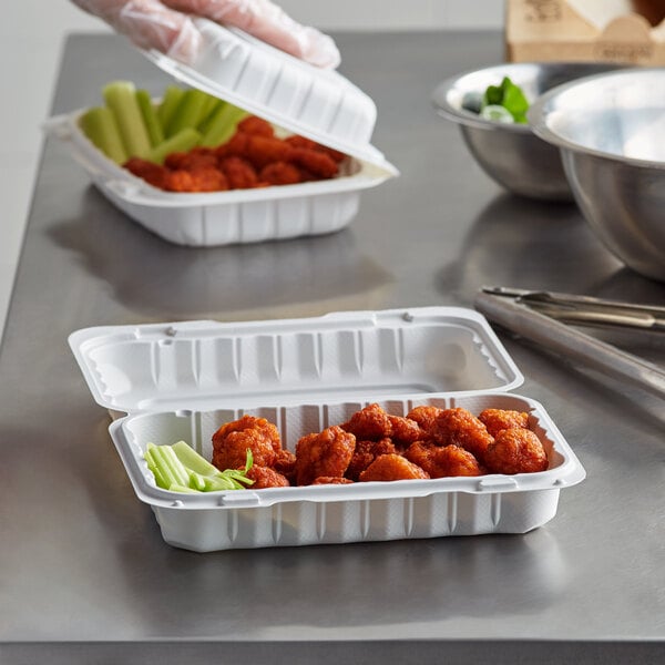 A person holding an Ecopax white plastic hinged container with chicken wings and celery on a counter.