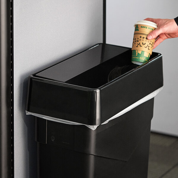A hand holding a paper coffee cup above a black trash can with a swing top lid.