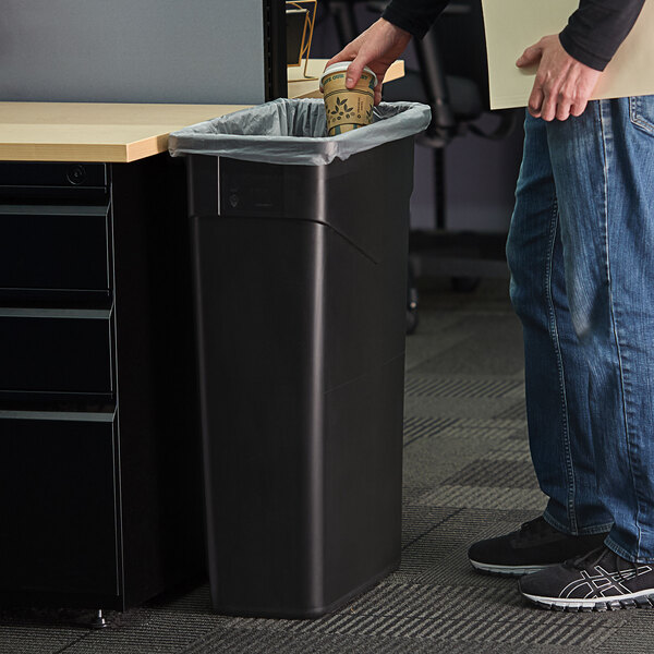 A man wearing jeans puts a coffee cup in a black Carlisle Trimline trash can.