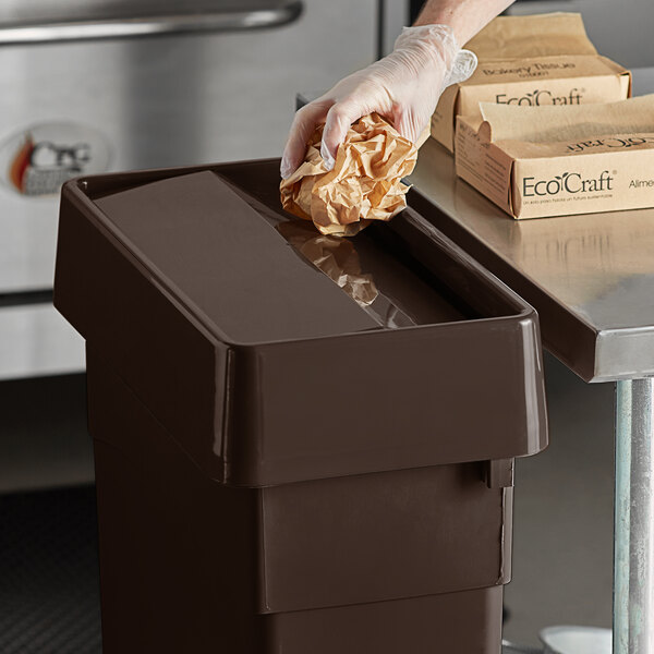 A hand in a glove throwing brown paper into a Carlisle brown trash can.