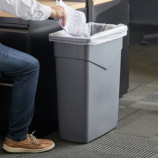 A person in a corporate office putting paper in a Carlisle grey rectangular trash can.