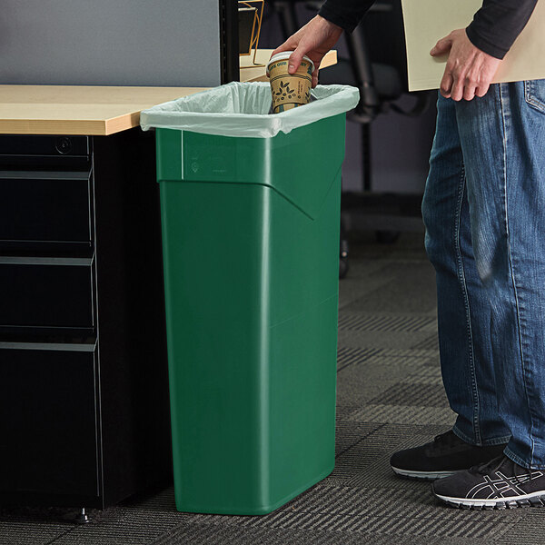 A person in a blue shirt standing next to a Carlisle green Trimline trash can putting a coffee cup in the trash.