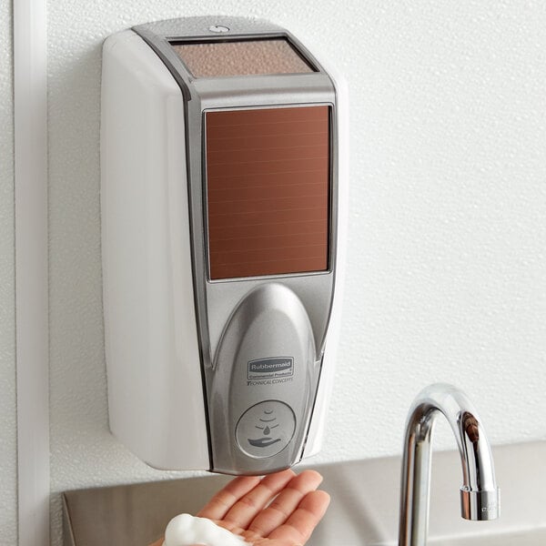 A person's hand using a white and grey Rubbermaid Lumecel automatic soap dispenser.