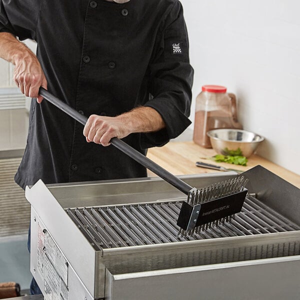 A man in a black chef coat using an American Metalcraft broiler brush with scraper on a grill.