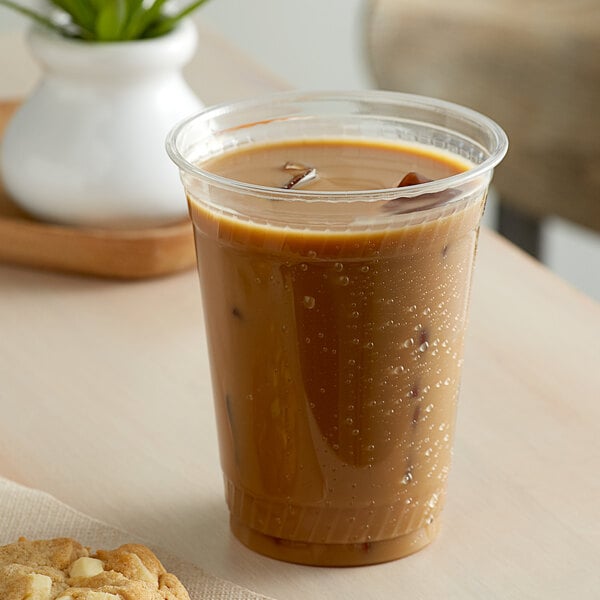 A clear Fabri-Kal plastic cup with brown liquid and ice cubes on a table with a cup of coffee and a cookie.