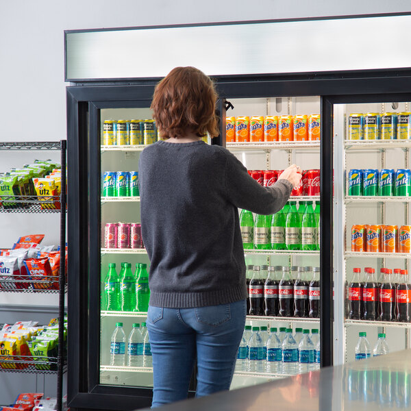A woman standing in front of a Beverage-Air glass sliding door refrigerator filled with drinks.