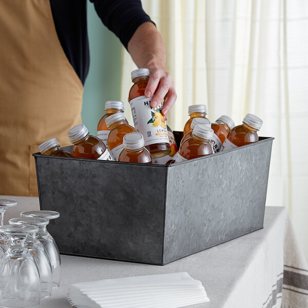 A person pouring juice into an American Metalcraft galvanized metal beverage tub filled with bottles of liquid.