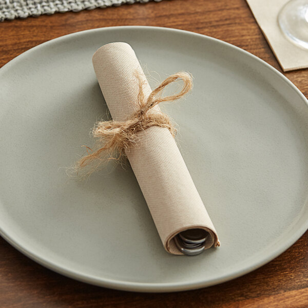 A place setting with a rolled up Hoffmaster Natural Dinner Napkin on a plate.