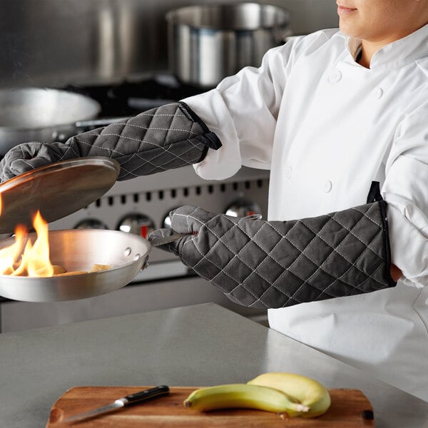 A chef holding a pan with a fire in it using San Jamar Bestguard oven mitts.
