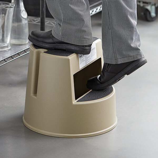 A person standing on a Rubbermaid Beige Mobile Two-Step Step Stool with black shoes.