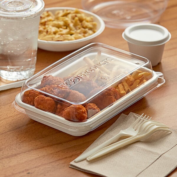 A white rectangular plastic container with a lid filled with food, chicken wings, and french fries on a table.
