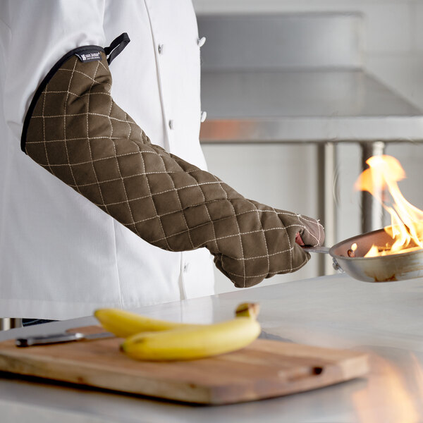 A person wearing a San Jamar brown oven mitt holding a pan over a flame.