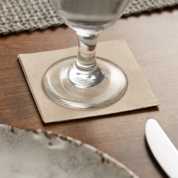A glass sits on a Hoffmaster FashnPoint natural beverage napkin on a table.