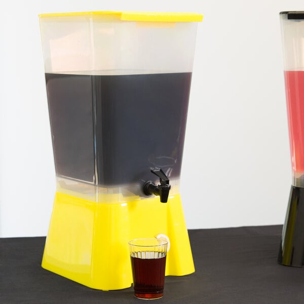 A yellow and black Tablecraft beverage dispenser with a drink in it and a glass of brown liquid.